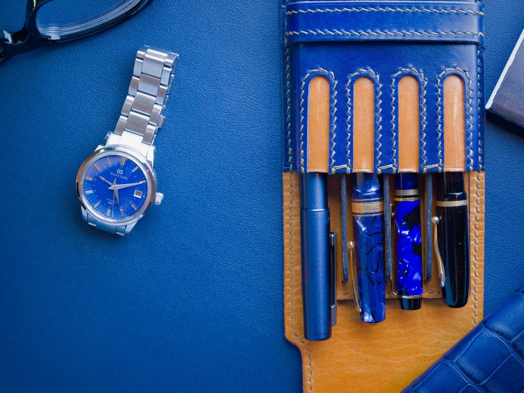 A blue leather pen case holding four fountain pens next to a silver watch with a blue dial and a pair of black glasses on a blue background.